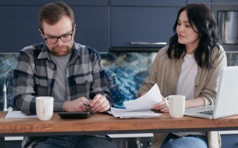 A couple discusses bills and finances using a laptop and calculator in their modern kitchen.