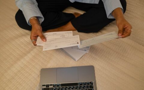 Close-up of hands holding cheques beside a laptop indoors for financial tasks.
