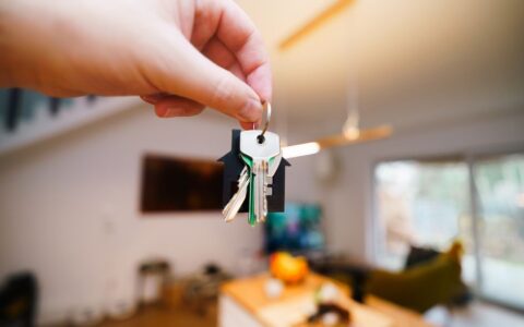 A hand holds keys with a house keychain inside a modern, well-lit living space.