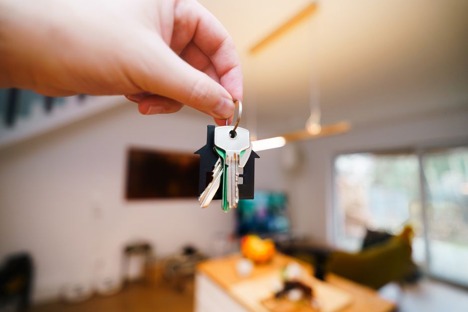 A hand holds keys with a house keychain inside a modern, well-lit living space.