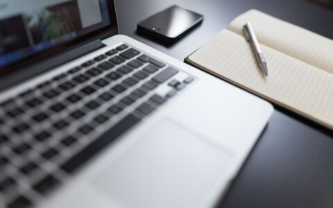 A modern workspace featuring a laptop, notebook, smartphone, and pen on a desk.