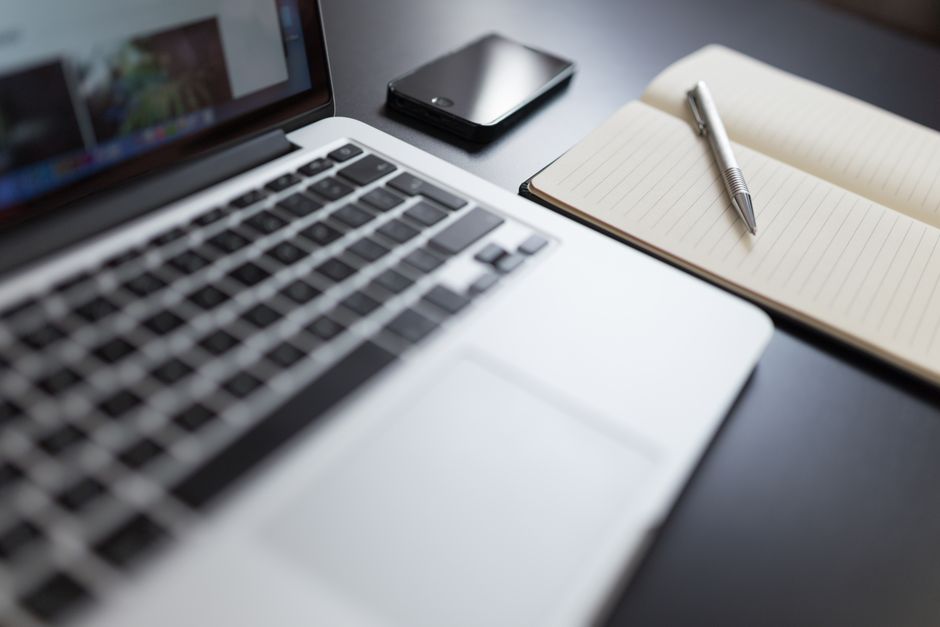 A modern workspace featuring a laptop, notebook, smartphone, and pen on a desk.