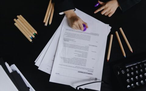 Hands working with documents and colored pencils on a black desk view.