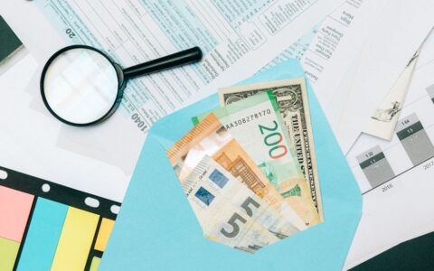 Close-up of tax documents and mixed currency in a blue envelope with a magnifying glass, perfect for financial analysis concepts.